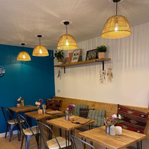 Cosy interior of The Coffee Hut in Helston, Cornwall, featuring stylish wicker lampshades and a soothing blue wall. The café's warm ambience is complemented by wooden tables, comfy bench seating with cushions, and charming décor, creating an inviting space for customers to enjoy their coffee and pastries.