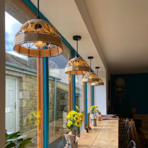 Interior view of The Coffee Hut in Helston, Cornwall, showcasing its rustic charm with wicker lampshades hanging above a wooden dining table. Sunlight streams through large windows illuminating the vibrant yellow daffodils and warm, welcoming atmosphere perfect for a relaxed coffee break."